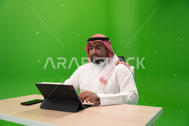 Completing work tasks via a tablet, using modern devices and technologies, a portrait of a young Saudi Arabian Gulf man wearing the traditional Saudi dress and shemagh sitting in his office, following up and managing administrative work, browsing websites