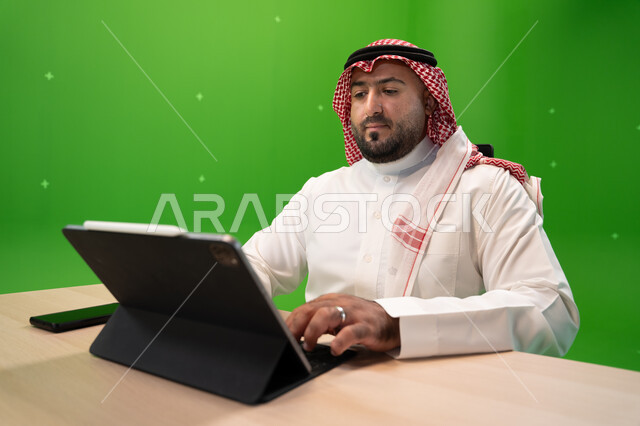 تصفح المواقع الالكترونية, استخدام الأجهزة والتقنيات الحديثة, بورتريه مقرب لشاب عربي خليجي سعودي يرتدي الثوب السعودي التقليدي والشماغ يجلس في مكتبه, إنجاز مهام العمل عبر جهاز التابليت, متابعة وإدارة الأعمال الادارية, خلفية خضراء, كروما