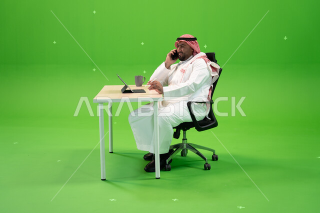 القيام بمكالمة هاتفية عبر الهاتف المحمول, بورتريه لموظف عربي سعودي خليجي يرتدي الثوب السعودي التقليدي والشماغ يقوم بالعمل داخل مكتبه, استخدام جهاز التابليت لإنجاز مهام العمل, إدارة شؤون الأعمال وتنظيمها, خلفية خضراء, كروما