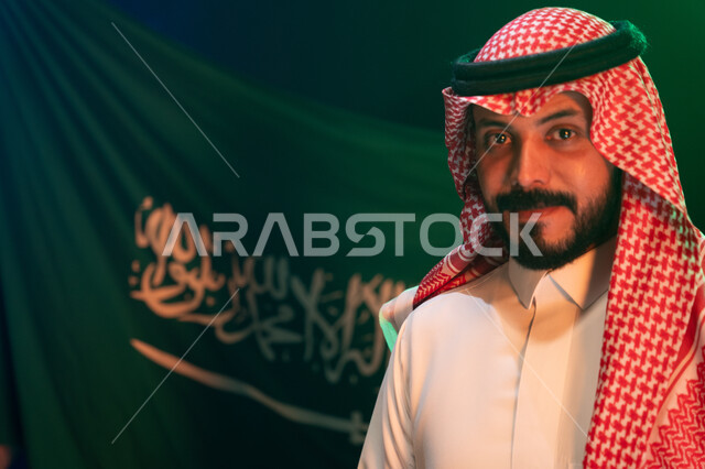 إظهار الحب والولاء للدولة, الاحتفال باليوم الوطني لتوحيد المملكة في 8 ربيع الأول, صوة مقربة لشاب عربي سعودي خليجي مبتسم يرتدي الثوب والشماغ التقليدي ينظر إلى الكاميرا بإيماءات الثقة, الوقوف أمام العلم في 11 مارس, خلفية العلم السعودي