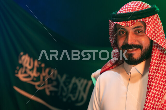 المشاركة في تنمية الوطن ورفع قدره, الاحتفال باليوم الوطني لتوحيد المملكة في 8 ربيع الأول, صوة مقربة لشاب عربي سعودي خليجي مبتسم يرتدي الثوب والشماغ التقليدي ينظر إلى الكاميرا بإيماءات الثقة, الوقوف أمام العلم في 11 مارس, خلفية العلم السعودي