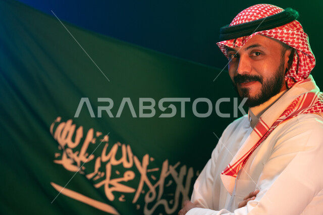 Looking at the camera with gestures of pride in Saudi identity, participating in national events on September 23 and remembering National Day, close-up photo from the side of a smiling Saudi Gulf Arab man wearing the traditional thobe and shemagh standing proudly on Flag Day March 11, showing loyalty to the state