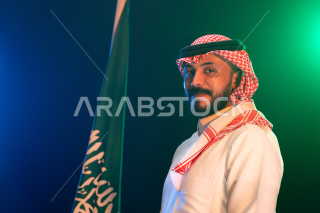 Participation in national events on September 23 and commemoration of National Day, pride in Saudi identity, close-up portrait of a smiling Saudi Gulf Arab man wearing traditional dress standing in front of the Kingdom’s flag with respect, commemoration of Saudi Flag Day March 11, blurry background with green lighting