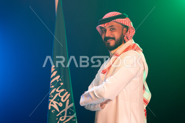 الفخر بالهوية السعودية, المشاركة في المناسبات الوطنية في 23 سبتمبر واستذكار اليوم الوطني, بورتريه مقرب من الجانب لرجل عربي سعودي خليجي مبتسم يرتدي الزي التقليدي يقف أمام علم المملكة باحترام, ذكرى يوم العلم السعودي 11 مارس, خلفية ضبابية مع إضاءة خضراء