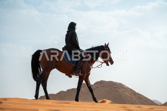 الفروسية وهواية ركوب الأحصنة في الصحراء، الاهتمام والشغف بركوب الجياد، صورة من الجانب لشابة عربية سعودية خليجية محجبة ترتدي العباءة تقوم بالتجول على ظهر جوادها العربي الأصيل في الصحراء، تعلق أبناء المملكة العربية السعودية بالخيول, أنشطة ترفيهية نسائية