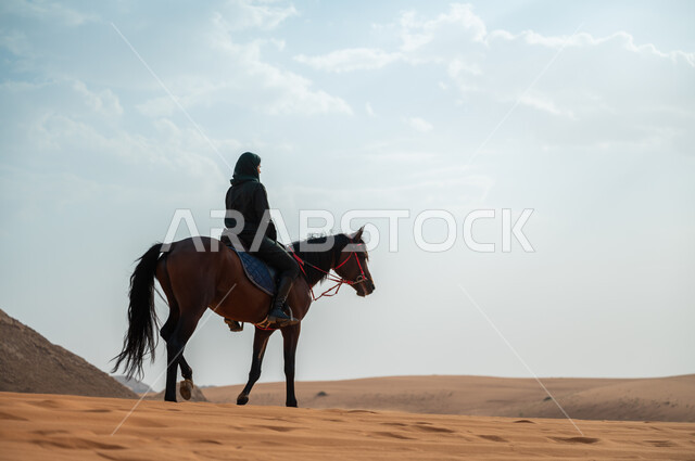 الفروسية وهواية ركوب الأحصنة في الصحراء، الاهتمام والشغف بركوب الجياد، صورة من الجانب لشابة عربية سعودية خليجية محجبة ترتدي العباءة تقوم بالتجول على ظهر جوادها العربي الأصيل في الصحراء، تعلق أبناء المملكة العربية السعودية بالخيول, أنشطة ترفيهية نسائية