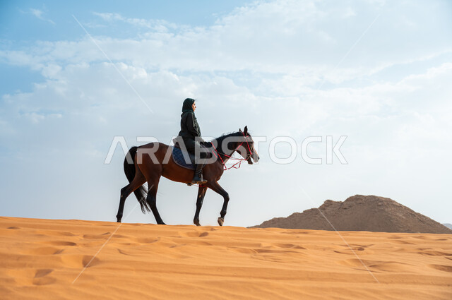  الاهتمام والشغف بركوب الجياد، الفروسية وهواية ركوب الأحصنة في الصحراء، صورة من الجانب لشابة عربية سعودية خليجية محجبة ترتدي العباءة تقوم بالتجول على ظهر جوادها العربي الأصيل في الصحراء، تعلق أبناء المملكة العربية السعودية بالخيول, أنشطة ترفيهية نسائية