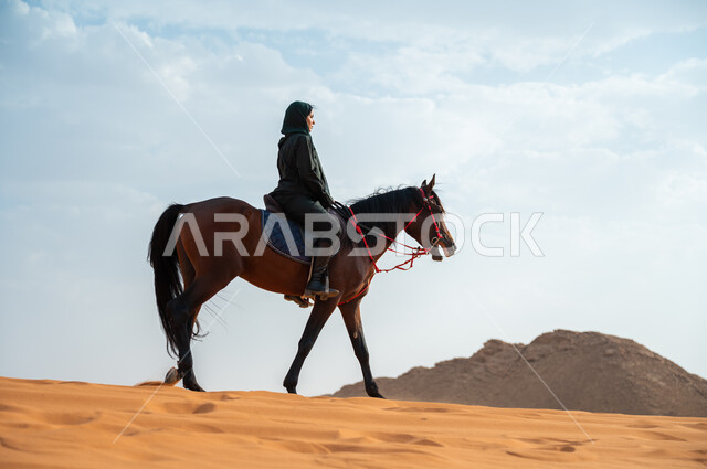 الاهتمام والشغف بركوب الجياد، الفروسية وهواية ركوب الأحصنة في الصحراء، صورة من الجانب لشابة عربية سعودية خليجية محجبة ترتدي العباءة تقوم بالتجول على ظهر جوادها العربي الأصيل في الصحراء، تعلق أبناء المملكة العربية السعودية بالخيول, أنشطة ترفيهية نسائية