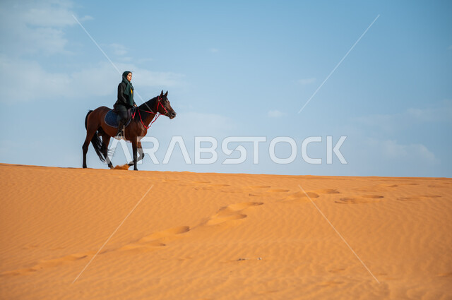 تعلق أبناء المملكة العربية السعودية بالخيول, الفروسية وهواية ركوب الأحصنة في الصحراء، الاهتمام والشغف بركوب الجياد، صورة من الجانب لشابة عربية سعودية خليجية محجبة ترتدي العباءة تقوم بالتجول على ظهر جوادها العربي الأصيل في الصحراء، أنشطة ترفيهية نسائية