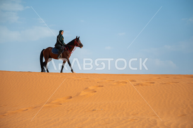 تعلق أبناء المملكة العربية السعودية بالخيول, الفروسية وهواية ركوب الأحصنة في الصحراء، الاهتمام والشغف بركوب الجياد، صورة من الجانب لشابة عربية سعودية خليجية محجبة ترتدي العباءة تقوم بالتجول على ظهر جوادها العربي الأصيل في الصحراء، أنشطة ترفيهية نسائية