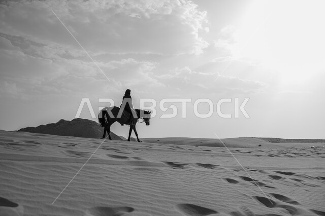 الفروسية وهواية ركوب الأحصنة في الصحراء، الاهتمام والشغف بركوب الجياد، صورة من الجانب بالأبيض و الأسود لشابة عربية سعودية خليجية محجبة ترتدي العباءة تقوم بالتجول على ظهر جوادها العربي الأصيل في الصحراء، تعلق أبناء المملكة العربية السعودية بالخيول