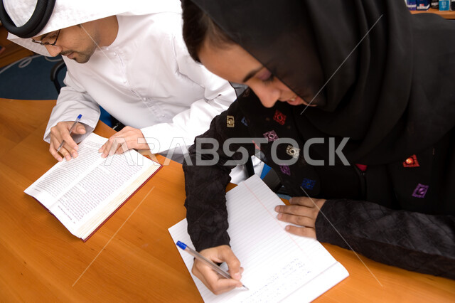 استخراج المعلومات وتدوينها وكتابة التقارير والأبحاث العلمية، صورة مقربة لطالب وطالبه عربيان خليجيان إماراتيان يقضيان الوقت بمراجعه الدروس اليومية في مكتبه الجامعه، ارتداء الزي الإماراتي التقليدي، الجامعات والكليات الإماراتية