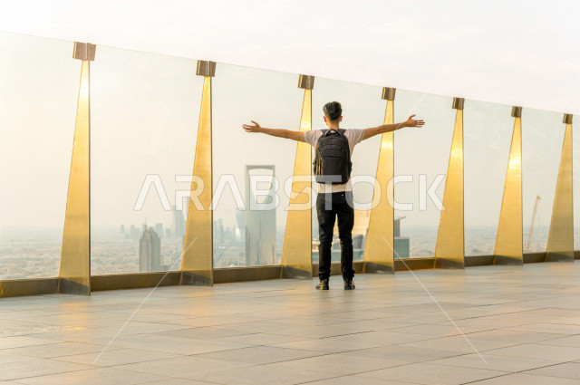 شاب سعودي سائح يحمل حقيبته على ظهره  ، يقف بشرفة عالية   ينظرالى ابراج الرياض ، اطلاله رائعة لأبراج الرياض ، ناطحات السحاب في المملكة العربية السعودية 