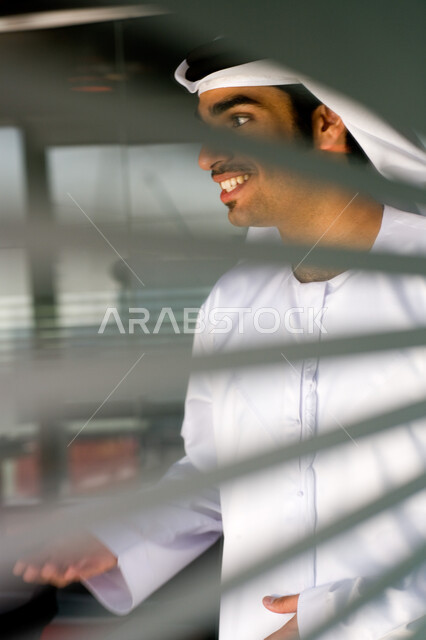 تبادل الأحاديث والنقاشات مع شركاء العمل، ايماءات السعادة والسرور، صورة مقربة لرجل عربي خليجي اماراتي يرتدي الغترة والكندورة يقف خلف جدار زجاجي مخطط في مقر الشركة، تحقيق إستراتيجيات وأهداف العمل ،مهن ووظائف في الامارات العربيه المتحدة