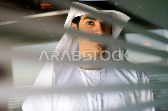 التأمل والنظر إلى الكاميرا، صورة مقربة لرجل عربي خليجي اماراتي يرتدي الغترة والكندورة يقف خلف جدار زجاجي مخطط في مقر الشركة، تحقيق إستراتيجيات وأهداف العمل ،مهن ووظائف في الامارات العربيه المتحدة