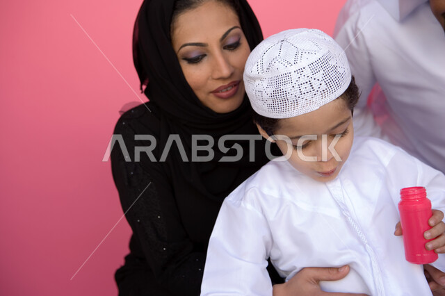 الاستمتاع بصنع فقاعة الصابون في الهواء, بورتريه مقرب لولد عربي خليجي إماراتي يرتدي الطاقية والكندورة الإماراتية يقوم بنفخ البالونات الهوائية برفقة والديه, اللعب بالفقاعات المائية المتطايرة والمتناثرة, تنمية مهارات الأطفال الحركية, خلفية وردية