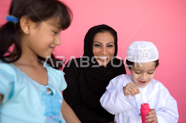 اللعب بالفقاعات المائية بإيماءات السعادة والفرح, قضاء الأوقات بالأنشطة الترفيهية الممتعه, بورتريه لعائلة عربية خليجية إماراتية تستمتع بصنع فقاعات الصابون, تنمية مهارات الأطفال الحركية, مفهوم الأسرة السعيدة, زرع روح المحبة بين الأبناء, خلفية وردية