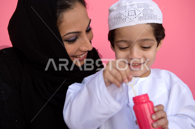 أهمية تنمية مهارات الأطفال الحركية, الاستمتاع بصنع فقاعة الصابون في الهواء, بورتريه مقرب لولد عربي خليجي إماراتي يرتدي الطاقية والكندورة الإماراتية يقوم بنفخ البالونات الهوائية برفقة والدته, اللعب بالفقاعات المائية المتطايرة والمتناثرة, خلفية وردية