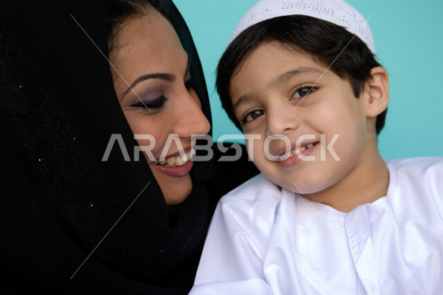 حنان الأم ومسؤوليتها تجاه أولادها, دور الأم في تربية أبنائها والعناية بهم, بورتريه مقرب لأم عربية إماراتية خليجية ترتدي العباءة والحجاب تنظر إلى طفلها بإيماءات الحب والحنان, مفهوم الطفولة والأمومة, قضاء أوقات ممتعة مع الأبناء, خلفية زرقاء