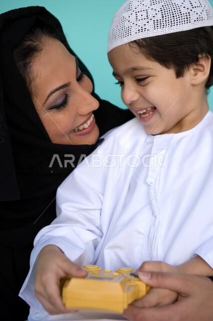 النظر إلى الطفل بإيماءات الحب والحنان, التسلية والترفيه بالفيديو جيمز, بورتريه مقرب لولد عربي خليجي إماراتي يرتدي الطاقية والكندورة يلعب في جهاز الألعاب المحمول برفقة والدته, الاستمتاع بالأنشطة الترفيهية الممتعة, تنمية قدرات الأطفال الذهنية, خلفية زرقاء