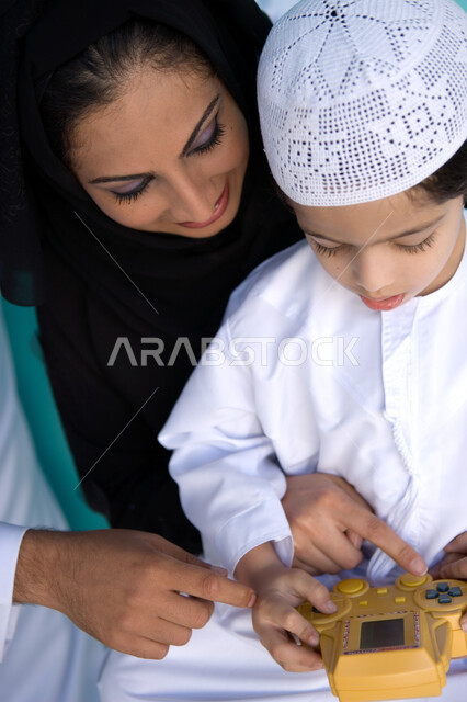 مشاركة الأبناء أوقات الفراغ, التسلية بالفيديو جيمز, بورتريه لولد عربي خليجي إماراتي يرتدي الزي الإماراتي التقليدي يلعب في جهاز الألعاب المحمول برفقة والدته, إيماءات السعادة بالأنشطة الترفيهية الممتعة, تنمية قدرات الأطفال الحركية والذهنية, خلفية زرقاء