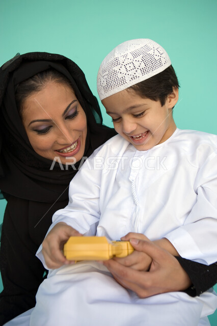 ايماءات التسلية والترفيه بالفيديو جيمز, بورتريه لولد عربي خليجي إماراتي يرتدي الكندورة يلعب في جهاز الألعاب المحمول برفقة والدته, الاستمتاع بالأنشطة الترفيهية الممتعة, أهمية مشاركة الأبناء أوقات الفراغ, تنمية قدرات الأطفال الحركية والذهنية, خلفية زرقاء