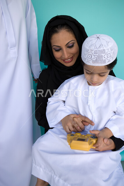 اللعب في جهاز الألعاب المحمول, التسلية والترفيه بالفيديو جيمز, بورتريه لولد عربي خليجي إماراتي يرتدي الكندورة والطاقية الإماراتية يجلس مع والديه بإيماءات السعادة, أهمية مشاركة الأبناء الأنشطة الترفيهية, تنمية قدرات الأطفال الحركية والذهنية, خلفية زرقاء