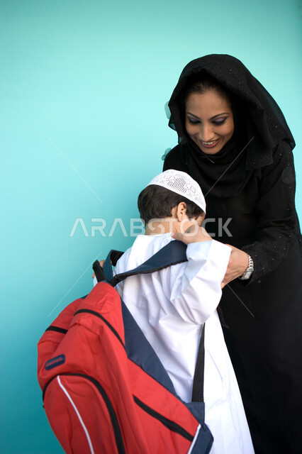 العودة إلى المدرسة, ارتداء الزي المدرسي الإماراتي, بورتريه لولد عربي خليجي إماراتي يرتدي حقيبة الظهر المدرسية, أم تساعد ابنها بتجهيز نفسه, الاستعداد للفصل الدراسي الجديد, دور الأم بالاهتمام بالأبناء, خلفية زرقاء
