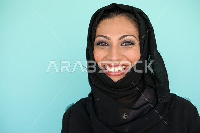 الاهتمام بالمظهر الخارجي, الوقوف بإيماءات السرور والسعادة, بورتريه مقرب لإمرأة عربية خليجية إماراتية مبتسمة ترتدي العباءة والحجاب تنظر إلى الكاميرا, الاستمتاع بقضاء وقت ممتع, نظرات الأنوثة الناعمة, خلفية زرقاء