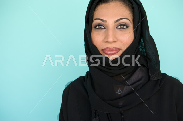 الوقوف بإيماءات السرور والسعادة, نظرات الأنوثة الناعمة, الاهتمام بالمظهر الخارجي, بورتريه مقرب لسيدة عربية خليجية إماراتية مبتسمة ترتدي العباءة والحجاب تنظر إلى الكاميرا, الاستمتاع بقضاء وقت ممتع, خلفية زرقاء