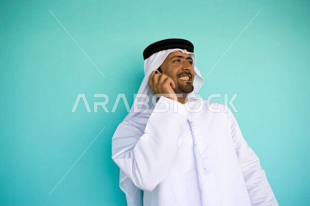 النظر إلى شيء ما وإجراء مكالمة هاتفية عبر الهاتف المحمول, بورتريه لشاب عربي خليجي إماراتي يرتدي الكندورة والغترة الإماراتية يقوم باستخدام الجوال, التواصل مع الأصدقاء بإيماءات السرور والسعادة, دردشات وأحاديث ودية عبر الانترنت, خلفية زرقاء