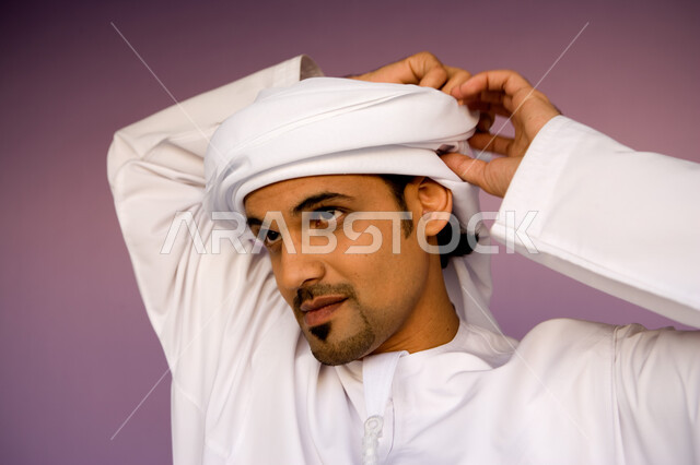 النظر إلى شيء ما بإيماءات التركيز, ارتداء الكندورة الاماراتية والعمامة, الاهتمام بالمظهر الخارجي, بورتريه مقرب لشاب عربي خليجي إماراتي يرتدي الزي التقليدي الإماراتي يقوم بلف العمامة, خلفية وردية