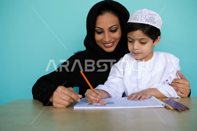 تنمية مهارات الأطفال ورعايتها، بورتريه لطفل عربي خليجي إماراتي يرتدي الزي الإماراتي يقوم بالرسم والتلوين برفقة والدته، التعلم وشغف حب الرسم، أم تساعد ابنها على الرسم بايماءات وجه تدل على السعادة والفخر، انشطة ترفيهية فنية ممتعه, خلفية زرقاء