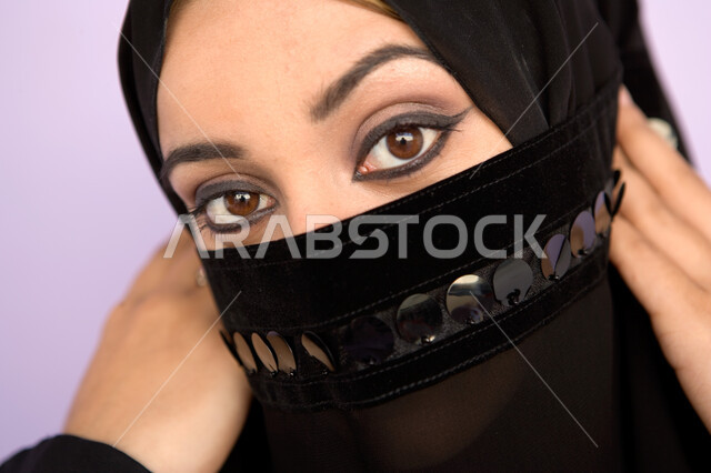 ابراز العيون المكحلة السوداء ، مفهوم الحياء والستر، بورتريه مقرب لوجه امراه عربية اماراتيه خليجيه محجبه تقوم بوضع اللثام على وجهها ، نظرات الأنوثه الناعمه، النظر الى الكاميرا، خلفية ملونه