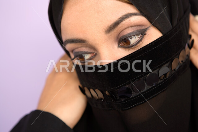النظر الى الأسفل لشيء ما، مفهوم الستر و الحياء، ابراز العيون المكحلة السوداء ، بورتريه مقرب لوجه امراه عربية اماراتيه خليجيه محجبه تقوم بوضع اللثام على وجهها ، نظرات الأنوثه الناعمه، خلفية ملونه