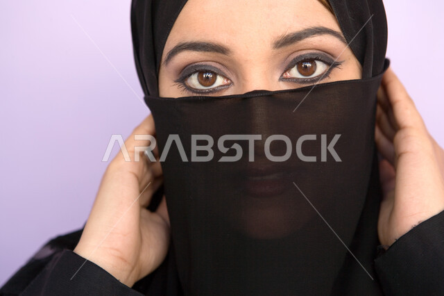 The concept of covering and modesty, highlighting the black kohl eyes, a close-up portrait of a veiled Arab-Emirati Gulf woman putting the veil on her face, soft feminine looks, looking at the camera, colored background.