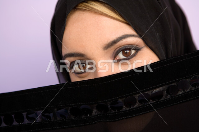 ابراز العيون المكحلة السوداء ، مفهوم الحياء والستر، بورتريه مقرب لوجه امراه عربية اماراتيه خليجيه محجبه تقوم بوضع اللثام على وجهها ، نظرات الأنوثه الناعمه، النظر الى الكاميرا، خلفية ملونه