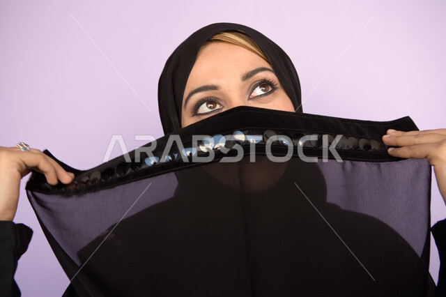مفهوم الحياء  والستر، بورتريه  مقرب لوجه امراه عربية اماراتيه خليجيه محجبه تقوم بوضع اللثام على وجهها ، ابراز العيون السوداء المكحله ، نظرات الأنوثه الناعمه، النظر الى الاعلى لشيء ما، خلفية ملونه