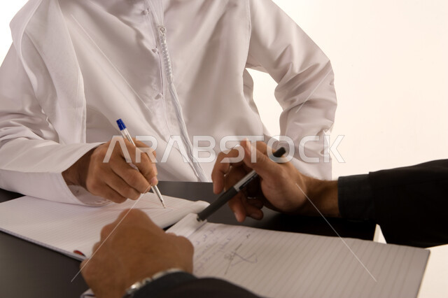 إعداد الخطط الاستراتيجية, إجراء المناقشات وتنظيم الأعمال, الشراكة والتعاون في انجاز العمل, صورة مقربة ليد رجل عربي خليجي اماراتي يرتدي الكندورة الاماراتية و يد رجل يتتبع المعلومات في غرفة الإجتماعات يقومان بتدوين الملاحظات والأفكار علبى دفتر الأعمال