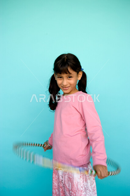  الاستمتاع في أوقات الفراغ والتسليه والترفيه، تعزيز المهارات الحركية للاطفال عن طريق اللعب, بورتريه لفتاة صغيرة عربية خليجية إماراتية تلعب بطوق الهولاهوب، النظر إلى الكاميرا بإيماءات السعادة، خلفية زرقاء