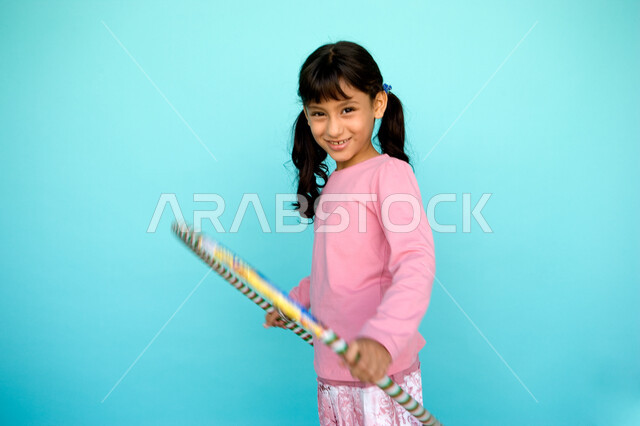 تعزيز المهارات الحركية للاطفال عن طريق اللعب, بورتريه لفتاة صغيرة عربية خليجية إماراتية تلعب بطوق الهولاهوب، النظر إلى الكاميرا بإيماءات السعادة، الاستمتاع في أوقات الفراغ والتسليه والترفيه، خلفية زرقاء