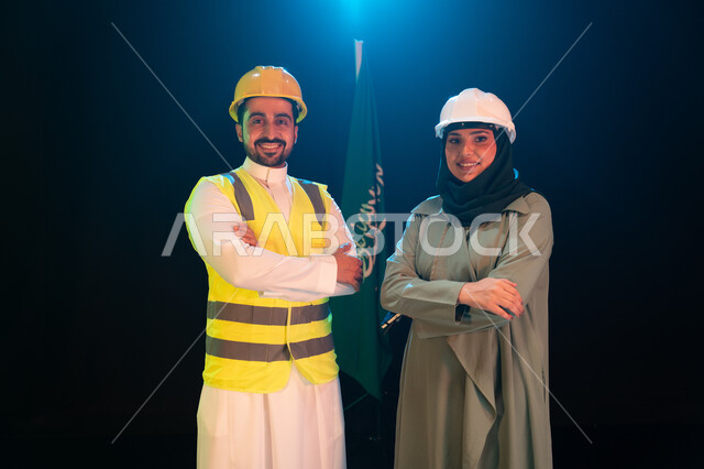 تطور القطاع الهندسي والإعمار بسواعد أبناء الوطن, صورة مقربة لمهندسان عربيان خليجيان سعوديان يرتديان خوذة الحماية يقفان بأيدي متقاطعة أمام علم السعودية، يوم العلم السعودي 11 مارس، الإحتفال بذكرى اليوم الوطني السعودي 23 سبتمبر، خلفية سوداء مع اضاءة بيضاء
