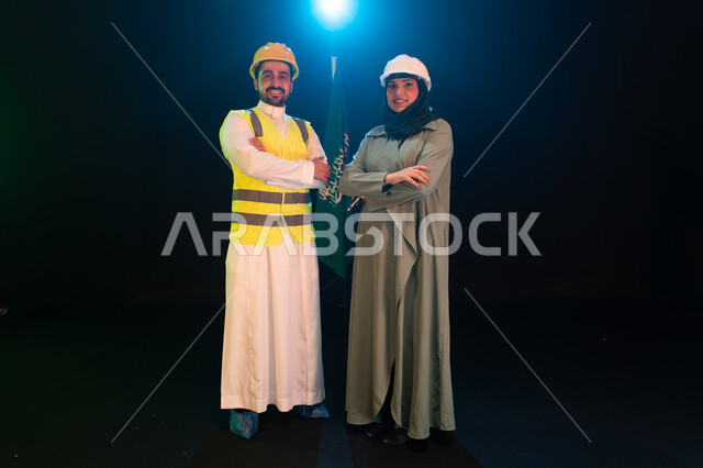 تنمية قطاع الهندسة والإعمار بسواعد أبناء الوطن, مهندسان عربيان خليجيان سعوديان يرتديان خوذة الحماية يقفان بأيدي متقاطعة أمام علم السعودية، يوم العلم 11مارس، الإحتفال بذكرى اليوم الوطني السعودي 23سبتمبر، صورة الجسم بالطول الكامل، خلفية سوداء مع اضاءة بيضاء