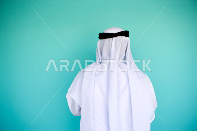 الوقوف باستقامة وثقة, الاهتمام بالمظهر الخارجي, بورتريه مقرب من الخلف لرجل عربي خليجي امارتي يرتدي الكندورة والغترة الاماراتية , خلفية زرقاء