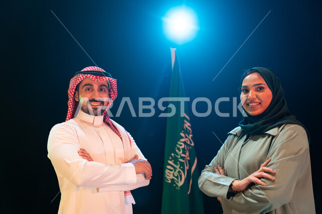 ايماءات الفخر والاعتزاز بالانتماء للوطن، الإحتفال بذكرى اليوم الوطني السعودي 23 سبتمبر، يوم العلم 11 مارس، صورة مقربة لرجل عربي خليجي سعودي يرتدي الشماغ والثوب التقليدي بجانبه إمرأة محجبة يقفان بأيدي متقاطعة امام علم السعودية، خلفية سوداء مع اضاءة بيضاء