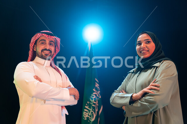 الإحتفال بذكرى اليوم الوطني السعودي 23 سبتمبر، يوم العلم 11 مارس، صورة مقربة لرجل عربي خليجي سعودي يرتدي الشماغ والثوب التقليدي بجانبه إمرأة محجبة يقفان بأيدي متقاطعة امام علم السعودية، ايماءات الفخر والاعتزاز بالانتماء للوطن، خلفية سوداء مع اضاءة بيضاء