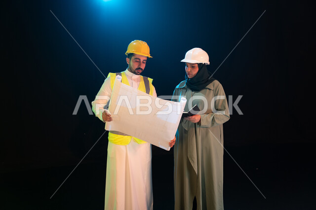 التدقيق والإشراف على العمل، مهن ووظائف سعودية، مفهوم المساواة بين الرجل والمرأة، مهندس عربي خليجي سعودي يقوم بالتعاون ومتابعة الاعمال والمخططات مع مهندسة محجبة، ارتداء سترة وخوذة الامان والحماية، خلفية سوداء مع اضاءة بيضاء.