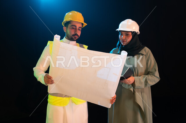مهن ووظائف سعودية، الإشراف والتدقيق على العمل، مفهوم المساواة بين الرجل والمرأة، صورة مقربة لمهندس عربي خليجي سعودي يقوم بالتعاون ومتابعة الاعمال والمخططات مع مهندسة محجبة، ارتداء سترة وخوذة الامان والحماية، خلفية سوداء مع اضاءة بيضاء.