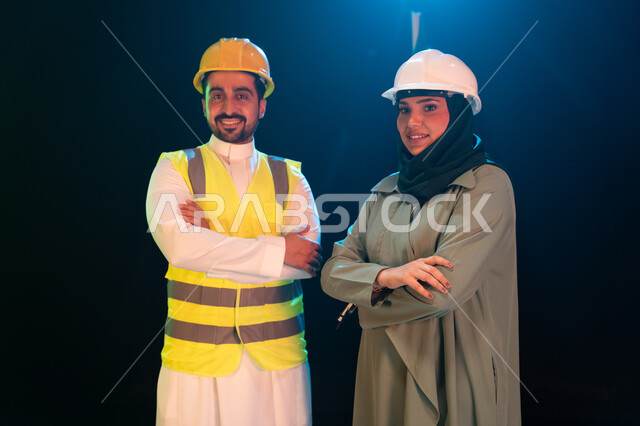  مفهوم المساواة بين الرجل والمرأة، ارتداء سترة وخوذة الامان والحماية، مهن ووظائف سعودية، بورتريه مقرب لمهندسان عربيان خليجيان سعوديان مبتسمان يقفان بثقة وأيدي متقاطعة، خلفية سوداء مع اضاءة بيضاء.