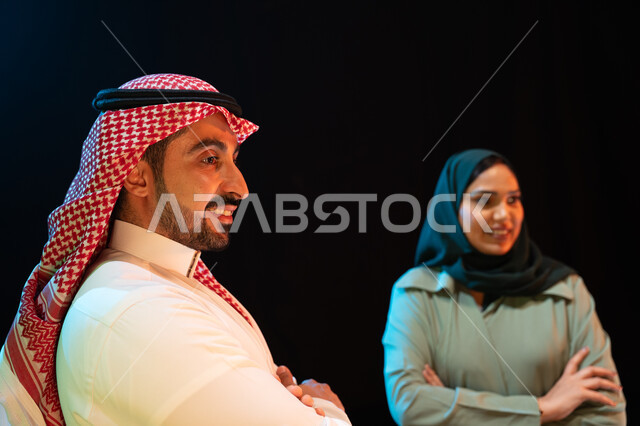 الوقوف بثقة وأيدي متقاطعه، بورتريه مقرب لإمرأة عربية خليجية سعودية محجبة تقف بجانب رجل يرتدي الشماغ والثوب السعودي التقليدي، مفهوم الترابط الاسري والعائلة السعيدة، ايماءات وجه تدل على السعادة والسرور، النظر الى شيء ما، خلفية سوداء